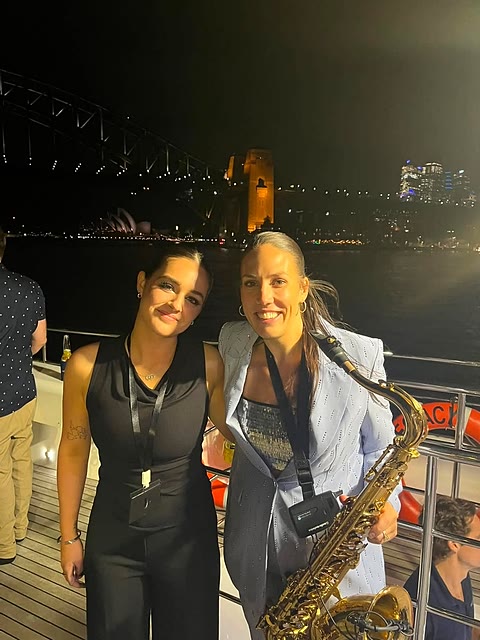 A guest and a live saxophonist on deck at night with the Sydney Harbour Bridge lit up behind them.